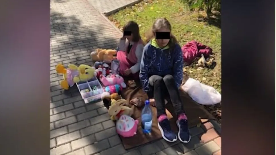 Două fetițe își vând jucăriile pe o pătură, în fața unui liceu